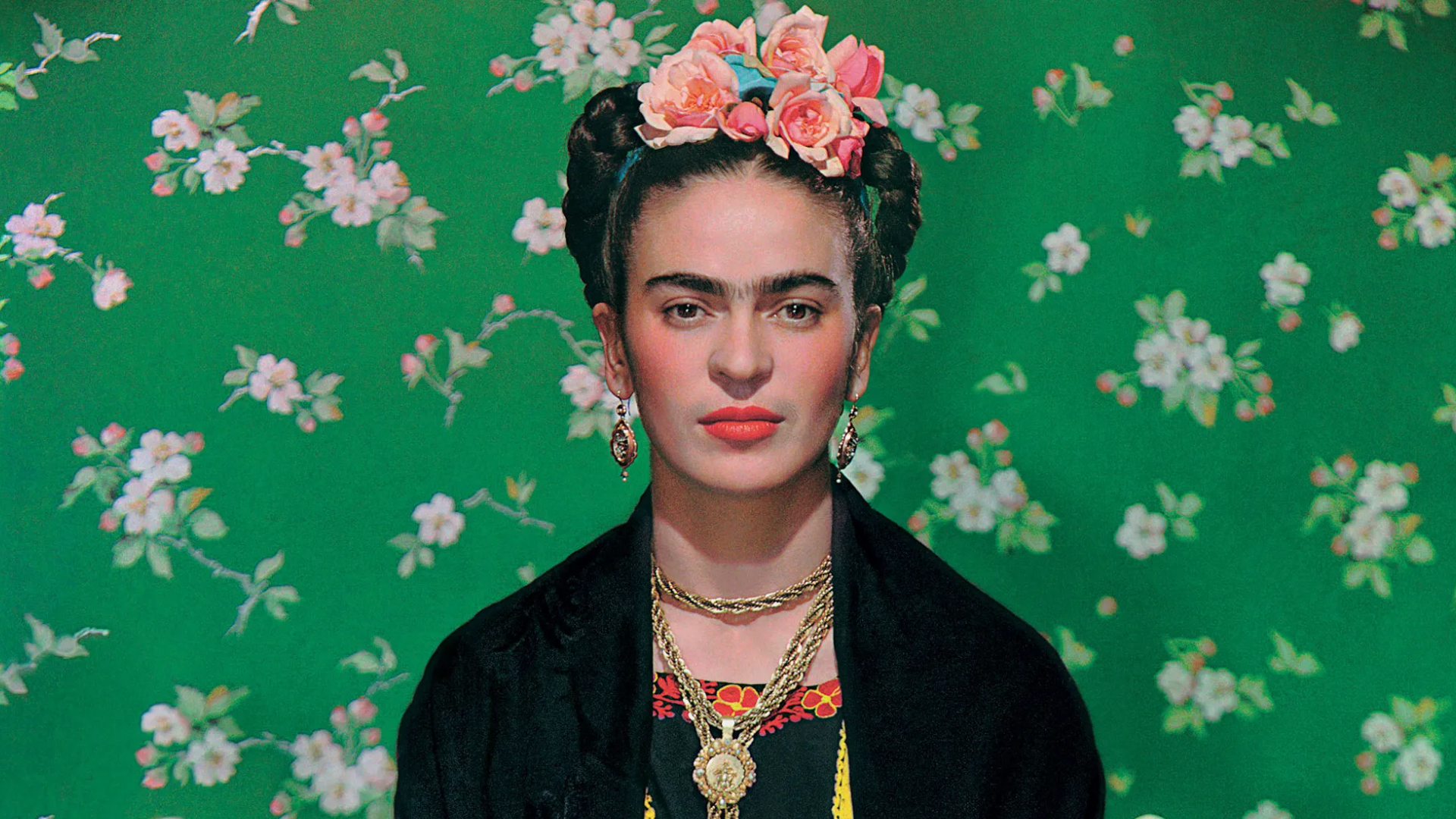 Frida Kahlo wearing luscious red lipstick, flowers in her hair, gold necklaces and long earrings, against a floral green background.