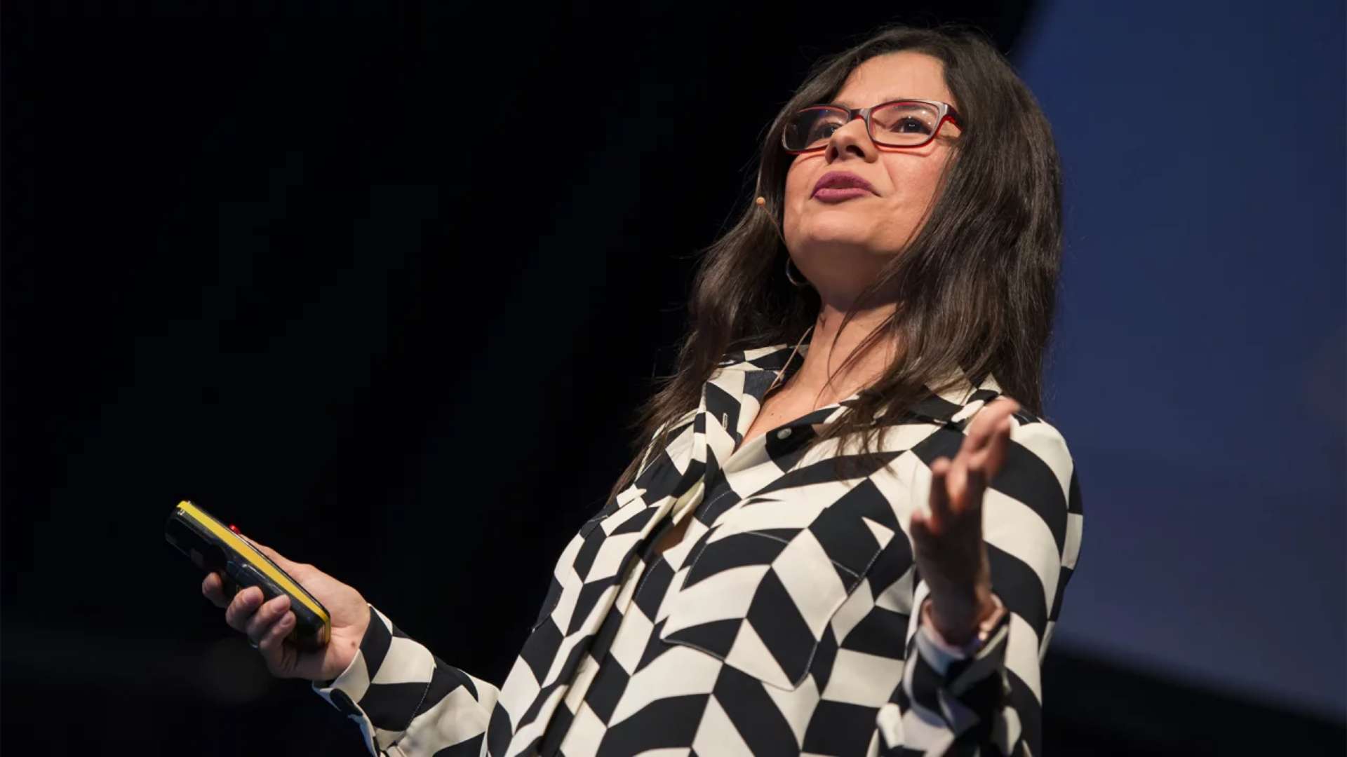 Aleyda Solís, wearing a black and white striped shirt and red framed glasses, standing on a stage doing a talk.