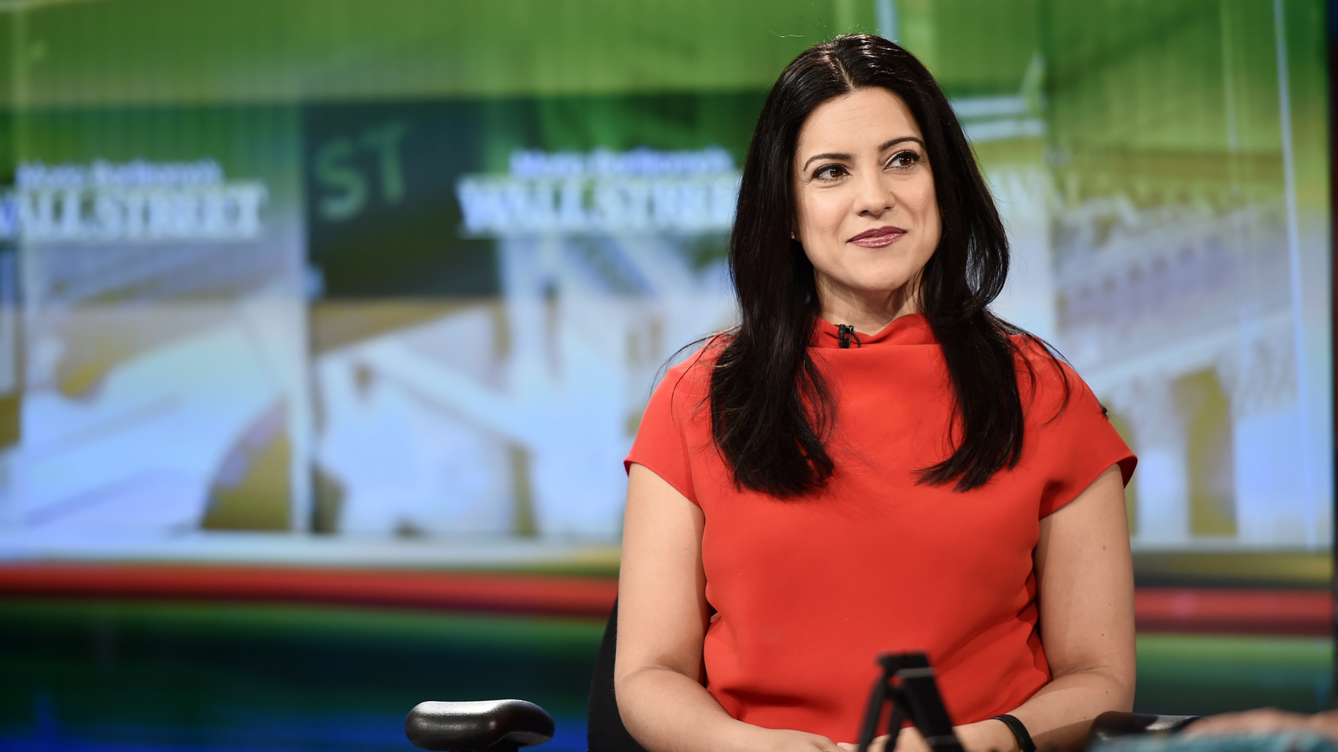 Reshma Saujani on a news show, wearing a red, short sleeved top, with long brown hair. There is a green news screen in the background.