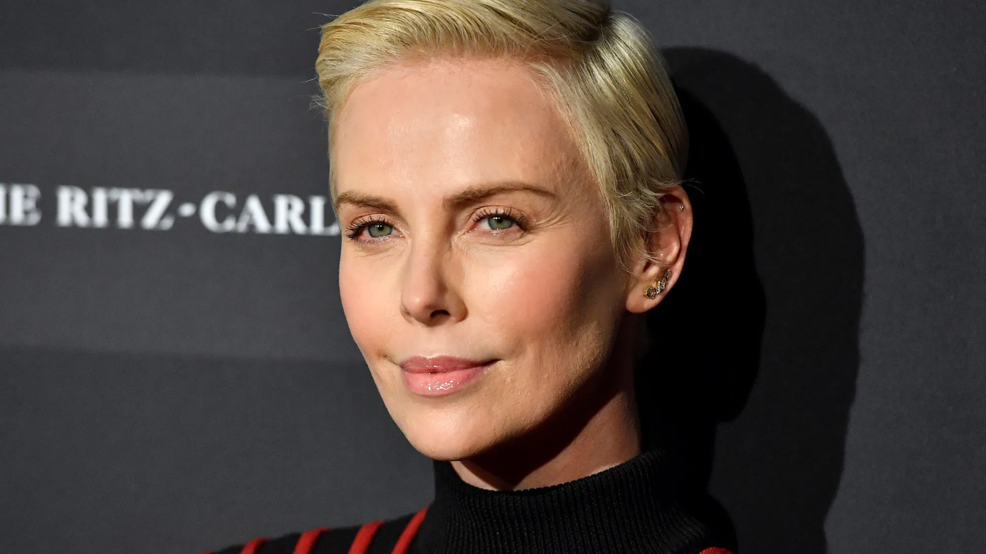 Charlize Theron on the red carpet, with a short blonde hair - wearing stripy black and red turtleneck jumper and stud earrings.