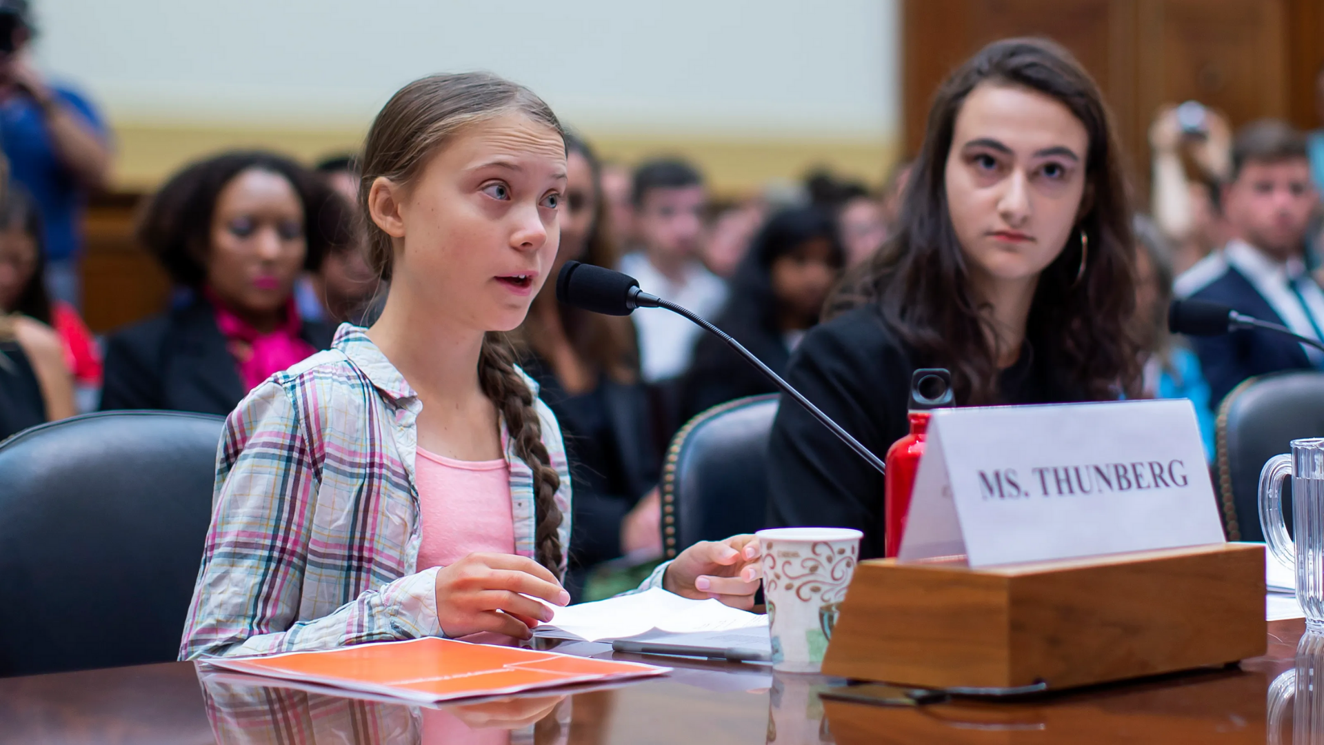 Greta Thunberg, sat talking into a microphone with a crowd of witnesses sat behind her.
