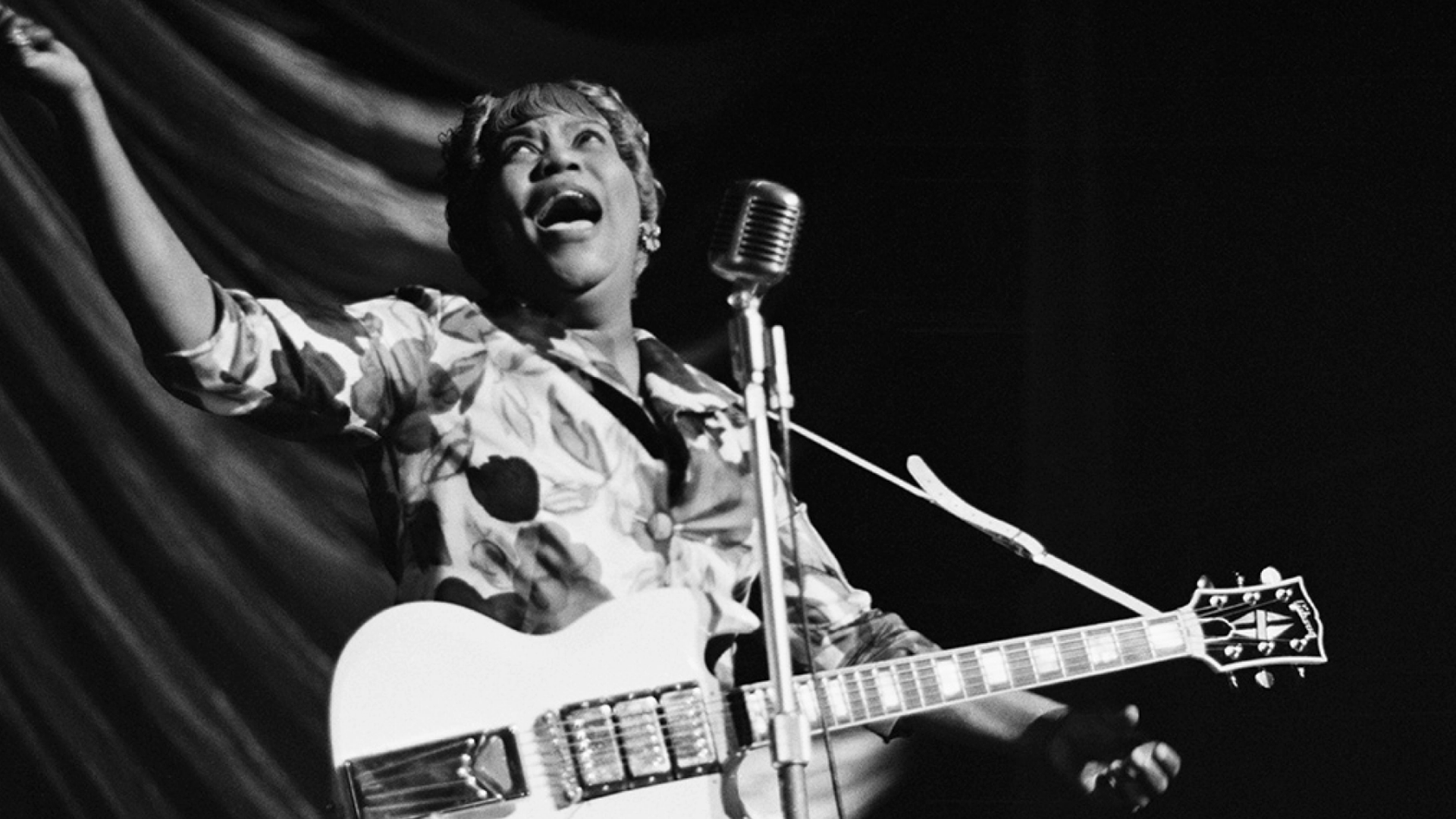 Old black and white photo of Sister Rosetta Tharpe, on stage with a guitar, singing into a microphone.